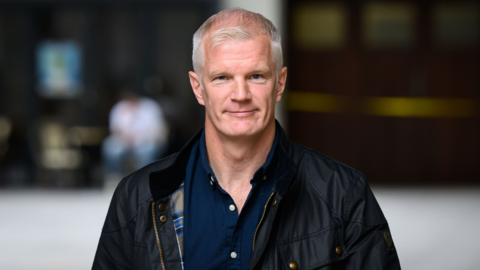 Al Carns is smiling. He has short grey hair. He's wearing an open navy shirt and a jacket. The background is blurred. 