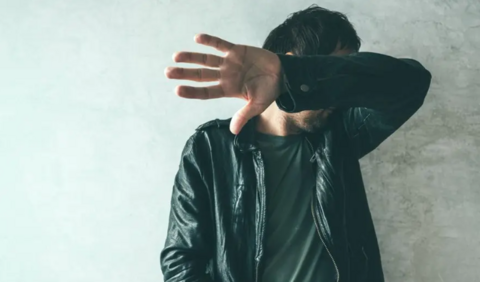 A man stands in front of a wall with his arm and hand up across his face, so we cannot see his facial features. He is wearing a black leather jacket and a t-shirt and has dark short hair.