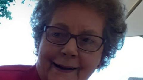 A picture of a woman smiling. She has grey hair and is wearing black glasses. She is wearing a red outfit. 