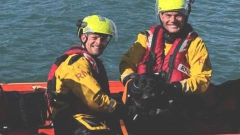 Two RNLI liveboat crew smile toward the camera wearing yellow uniforms with red life jackets in a boat and holding on to a black dog.