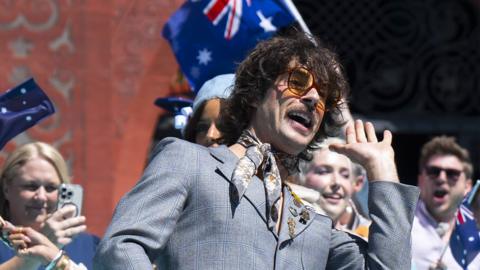 Australia's Go-Jo, in a grey suit, poses with an Australian flag on the Eurovision turquoise carpet