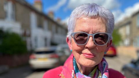 Head and shoulders image of Tessa Byers standing in the middle of her street, with houses and cars on both sides of her. Tessa has short white hair, is wearing rounded sunglasses, a pink top and multi-coloured scarf.