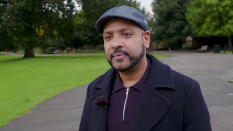 A man in a purple collared shirt, a navy blue coat, a blue flat cap. He has a beard and moustache. He stands in a park, with trees and grass in the background.