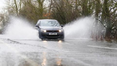 A dark grey sedan car with full beams on sprays standing water either side of it as it goes down a wet, tree-lined road.