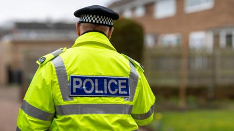 A stock photo of the back of a police officer wearing a bright yellow jacket with the words "police" printed on his back.