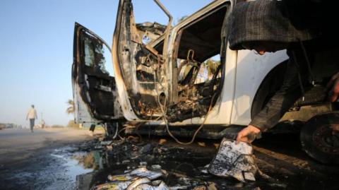 A Palestinian in a hooded jumper crouches beside a destroyed vehicle, with debris and scorched World Central Kitchen tags on the ground.