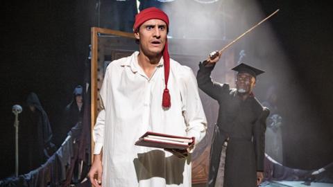 A male actor is wearing Victorian nightgown performing as a character on stage with another male actor in a Victorian teacher's costume and a cane in his hand standing by a chalk board.