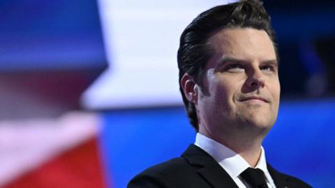 Matt Gaetz in a suit standing in front of a a blue, white and red background