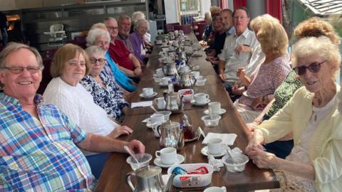 A mixed group of older people sit around a wooden table. The table has plates and mugs on it, and in the background other tables with groups of people sat around can also be seen.