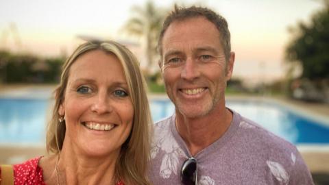 Lindsay Foreman and Craig Foreman smile as they pose together for a selfie picture. Behind them is a swimming pool. She is wearing a red top and him a lilac T-shirt.