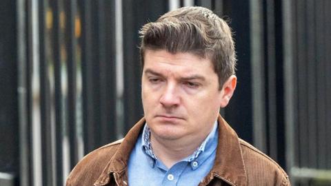 Michael McMonagle walking into court wearing a blue shirt and brown jacket. His hair is short and brown. The courthouse railings can be seen in the background