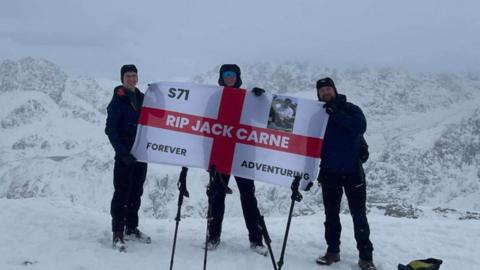Matty Belcher, Brandan Smith and Jack's dad, Rich, they are all holding the England flag that has 'rip Jack Carne' in capitals on it and also 'forever adventuring' as well. They are standing in front of snowy white mountains. 