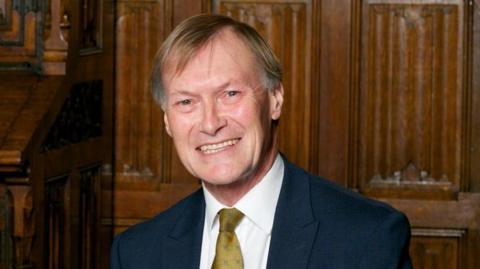 Sir David Amess, wearing a dark suit, white shirt and gold-coloured tie, smiles at the camera. He is seated. Behind him is a wood-panelled wall.