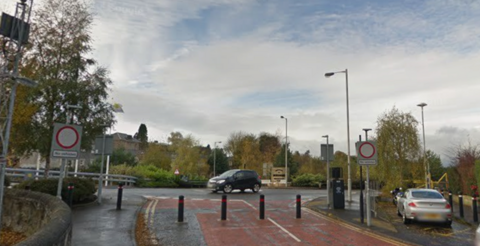 A roundabout with the road to the south of the photo pedestrianised with bollards. Parked cars to the right and a car is travelling through the roundabout