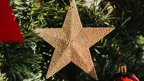 A gold star decoration hanging on the branch of a green Christmas tree, other red baubles can be glimpsed at the edges