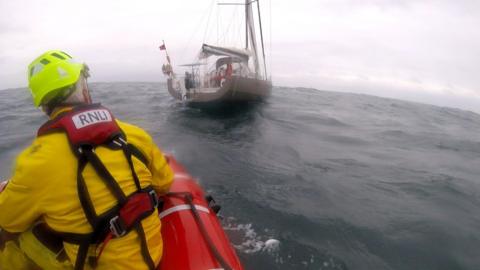An RNLI rescuer wearing water safety equipment sitting on the edge of a lifeboat looking towards a yacht in the ocean. The skies are grey.