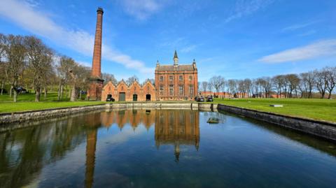 View of a Victorian building - a former pumping station - in red brick with two high storeys and with tall windows, and a pitched roof with a small turret in the middle. To its side is a line of four brick buildings with pitched roofs and a separate high chimney. The scene is reflected in a body of water in front, with a line of leafless trees on either side under blue skies.
