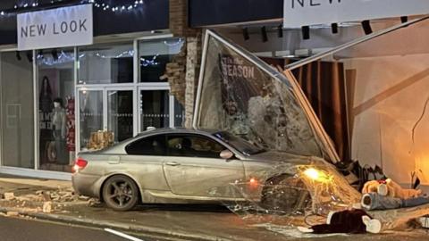 A grey saloon-style vehicle that has crashed into the front of a New Look shop. The car's lights are still on and the shop window has smashed around it. Debris can be seen on the ground. 