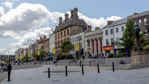 Wide view of a pedestrian-friendly town centre street lined with historic and modern buildings under a bright blue sky with large white clouds. Stone steps lead up to shops and businesses. A statue stands on a raised plinth at the right side of the square. Streetlights, trees, bollards, and scattered pedestrians are visible along the gently curving paved roadway and public plaza.