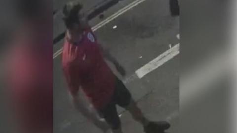 A CCTV image of a man, taken from above, from the street. He is standing in the middle of the road, and is wearing a red, short-sleeved England shirt. He is wearing dark shorts and shoes, has dark hair, and his face is not clearly visible due to being turned away. 