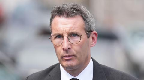  Muir is outside. He's wearing a black suit with a white short and a black tie. 