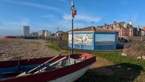 The rotunda at Windsor Lawns in Worthing, West Sussex, which has been boarded up since 2021 following a spate of antisocial behaviour. To the left of the rotunda is a pebbled beach and a red row boat.