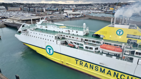 An aerial view of a white and yellow boat arriving into St Helier harbour. You can see rows of built up buildings and roads in the distance. Grey skies.