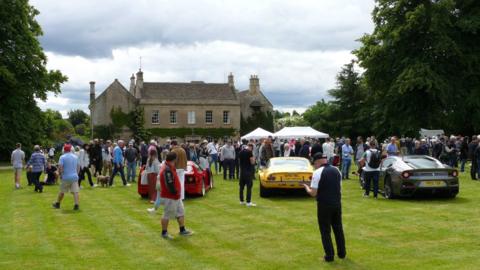 A large number of people stand on a lawn outside a house. There are three classic cars parked on the grass.