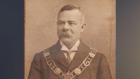 Allan Minns in about 1904, in a sepia-toned black and white photo. He is has short greying hair and a large bristling moustache, covering his upper lip. He is wearing a jacket and underneath a wing-tipped white shirt. Over his jacket is a mayoral chain. It has two rows of links and a large medallion dangling across his stomach