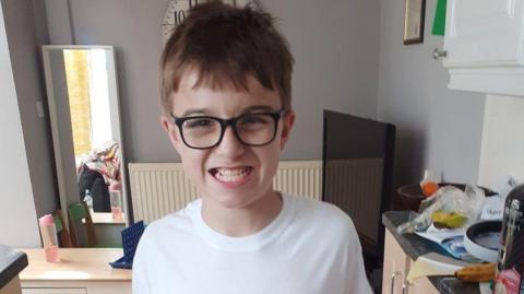 Twelve-year-old Lucas, pictured inside his home,  grinning widely at the camera, wearing glasses and a white T-shirt