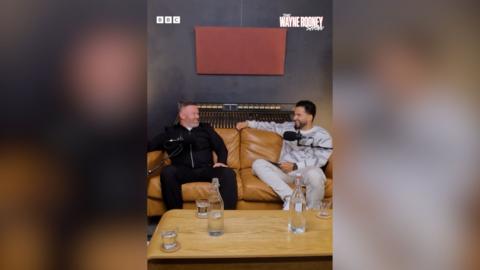 Two men, one wearing a black tracksuit, one wearing a grey one, sit side by side on a brown sofa