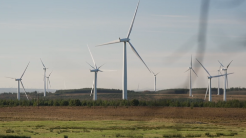 About 10 white wind turbines spinning on a green and brown field site with a grey sky overheard
