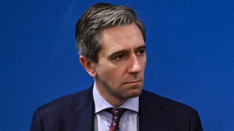 A close up of Simon Harris  He has short, greying hair and is wearing a navy suit, a lilac shirt and a maroon patterned tie