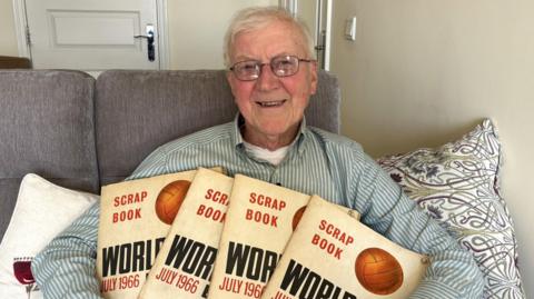 A man holding 4 scrapbooks while smiling into the camera he is wearing a stripey green and white top and has white hair and glasses.