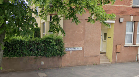 A sign reading WITHYCOMBE ROAD is mounted on the brown wall of a house along the road. There is a tree and hedge to the left and a yellow doorway to the right. 