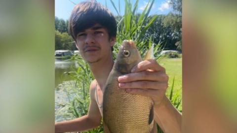 Holton Knott looking at the camera holding up a fish. He is sitting next to a riverbank.