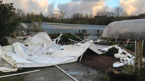 A white marquee is upside down and all the poles are twisted and broken, behind that is a poly tunnel which has remained unscathed and a 30 metre long glasshouse that has panes missing in it. 