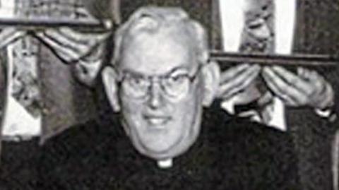 A black and white photo of the late Fr Malachy Finegan. The priest had short white hair and glasses and wore a black suit with a white clerical collar. 