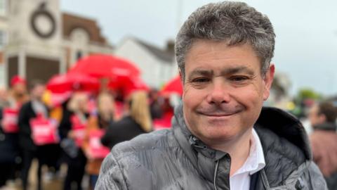 John Kent smiling at the camera while wearing a grey coat over a white shirt. It is a rainy day and he is standing in front of Labour campaigners with red umbrellas and placards