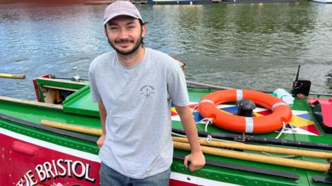 Jude Taljaard leans against a green and red narrowboat that is moored in a harbour. He wears a grey Bristol Packet tee-shirt and baseball cap. 