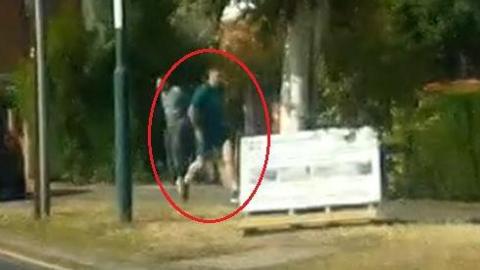 A very low quality image of a man running or walking down a street wearing a green T-shirt and shorts. 