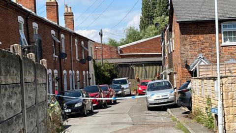 Police tape has been put up across a residential street where a number of cars are parked.