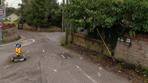 One entry to Dark lane, with the road sign showing on the right of the picture. There is a bollard in the road and an accept for access street sign to the left. There are trees in the picture, around, and above, walls as you enter the road. 