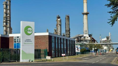 The entrance to an oil refinery with a large white and green sign, reading "Prax Lindsey Oil Refinery Main Entrance" in front of a two-storey red-brick and glass building. A road leads towards tall metallic chimneys and other refinery structures under a blue sky.