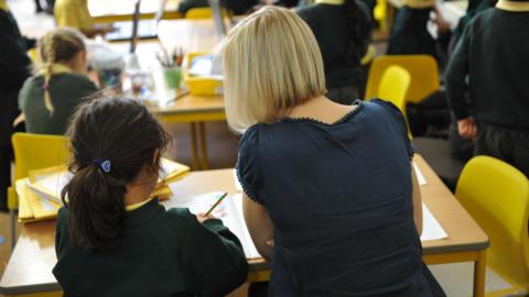 A stock image  - a female teacher with short blonde hair and a dark blue short-sleeved top helps an anonymous young girl of primary school age, at a classroom desk with yellow chairs. The pupil is wearing a dark green jumper and has a blue toggle with a heart on it in her tied up brown hair.