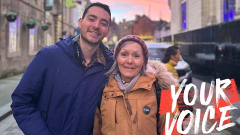Deborah and her son Stuart smile while wearing coats - Stuart is wearing a royal blue one and hers is a burnt orange. They are standing in the street, with a pink sunset in the sky behind them. In the bottom right corner of the image is a white and red 'Your Voice' logo.