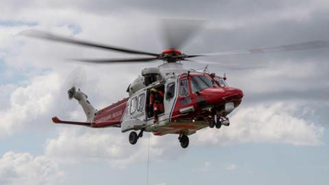 A red and white HM Coastguard helicopter is in the air. A rescuer holding a rescue rope out of the side of the helicopter.