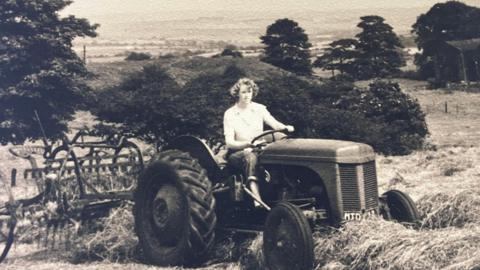 A black-and-white photograph of Margaret Lees taken in 1955. She is sitting on a tractor in a field full of hay. 