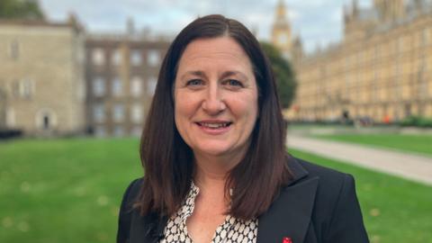 MP Julia Buckley standing in front of the Palace of Westminster wearing a black suit and checkered blouse.