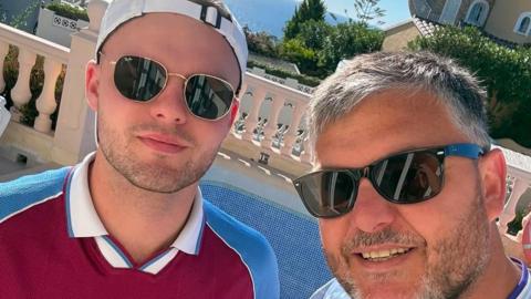 Two men wearing sunglasses are looking at the camera. One is wearing a Villa shirt with a white cap on backwards and stubble. The other has a beard and grey hair. The sun is shining and there is greenery behind.